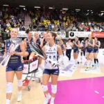 Atletas de voleibol feminino exibem troféus após vitória em partida disputada em ginásio com arquibancadas repletas de torcedores