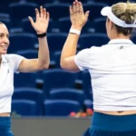 Duas tenistas de uniforme branco e saia azul comemoram um ponto com um high five em quadra de estádio com arquibancadas vazias em azul