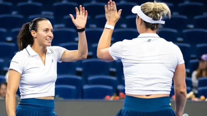 Duas tenistas de uniforme branco e saia azul comemoram um ponto com um high five em quadra de estádio com arquibancadas vazias em azul