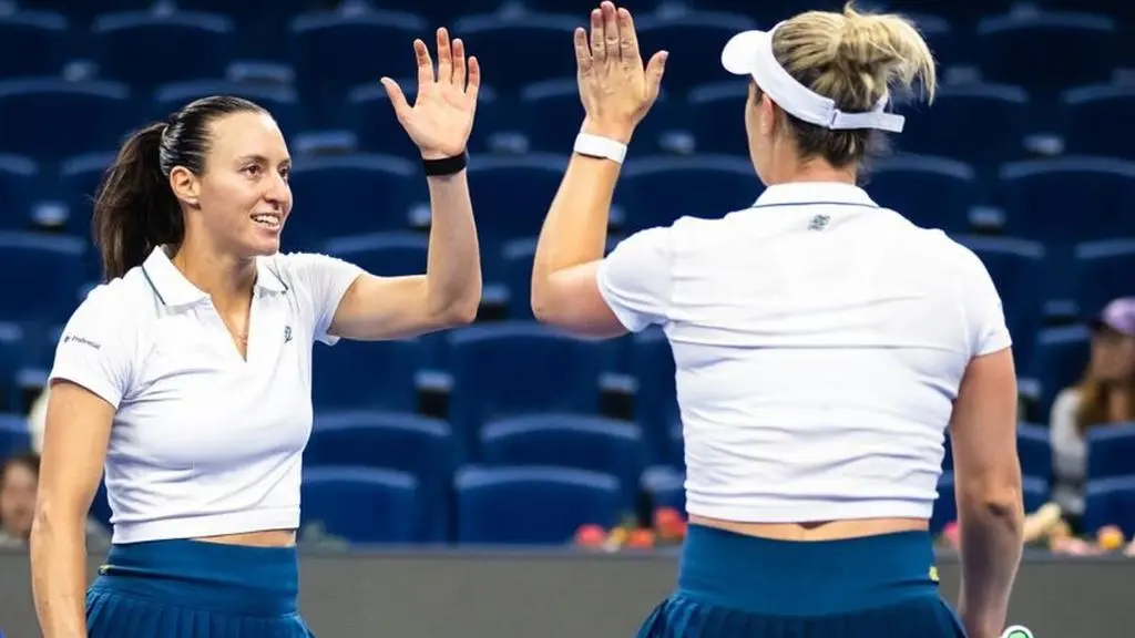 Duas tenistas de uniforme branco e saia azul comemoram um ponto com um high five em quadra de estádio com arquibancadas vazias em azul