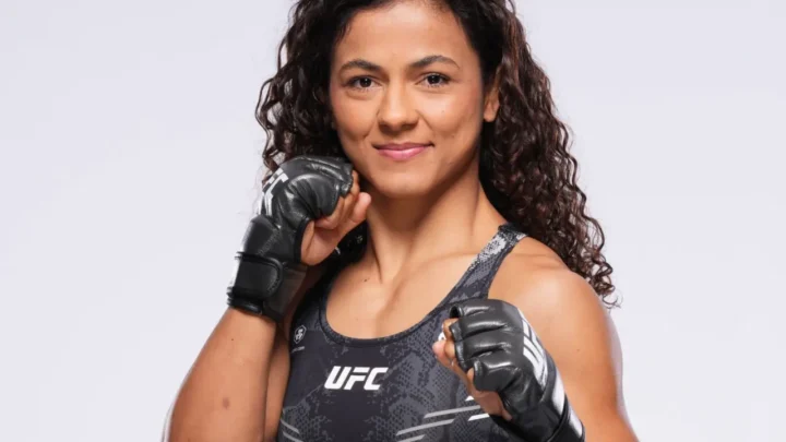 Mulher de cabelo comprido ondulado em uniforme de competição UFC, usando luvas de combate pretas, em pose frontal contra fundo cinzento