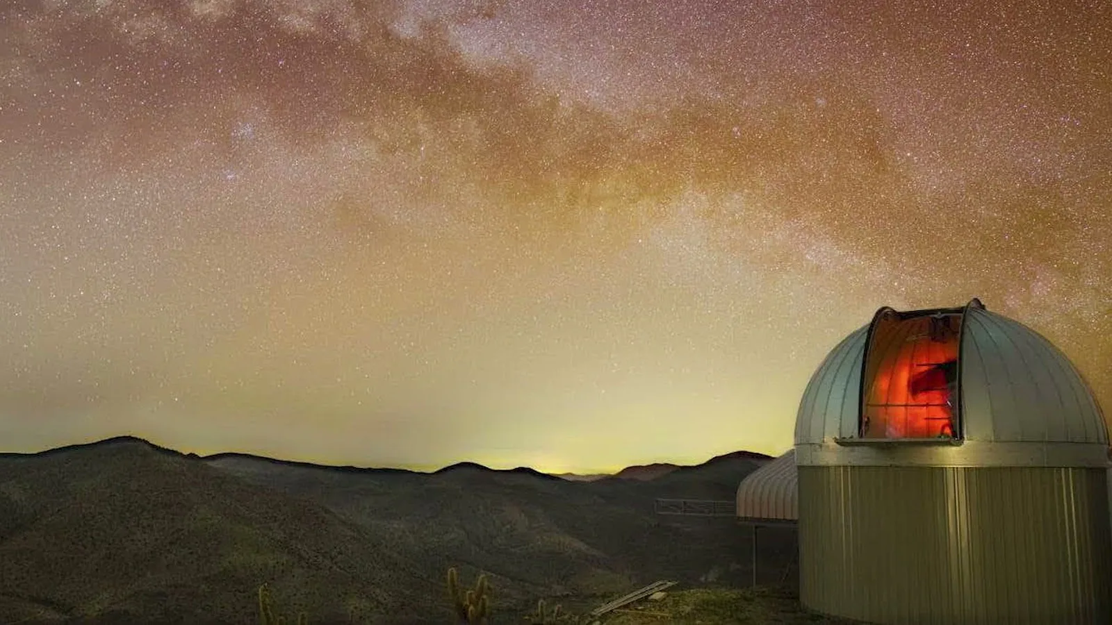 Cúpula de observatório aberta, com luz vermelha no interior, sob a Via Láctea em uma montanha à noite