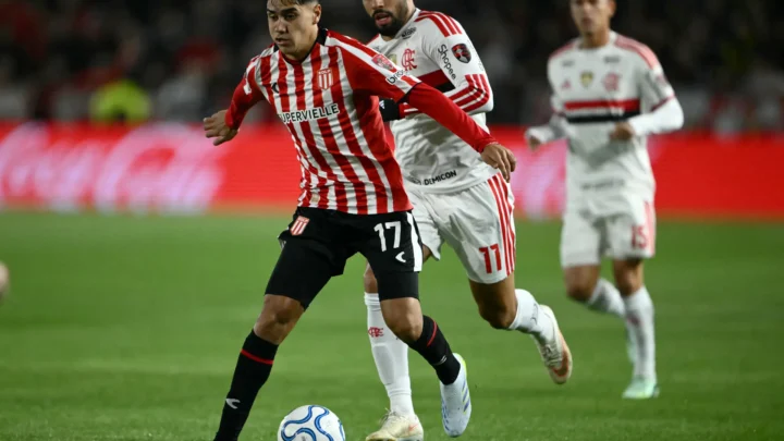 Jogador do Estudiantes, de camisa vermelha e branca listrada e número 17, conduz a bola sob marcação de um atleta do Flamengo, de uniforme branco com listras vermelhas e número 11