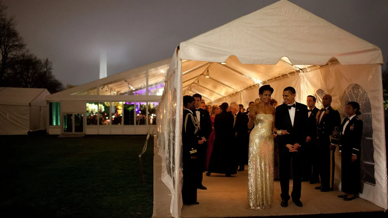 Casal em evento noturno dentro de tenda iluminada, cercado por convidados em trajes formais, com edifício iluminado ao fundo