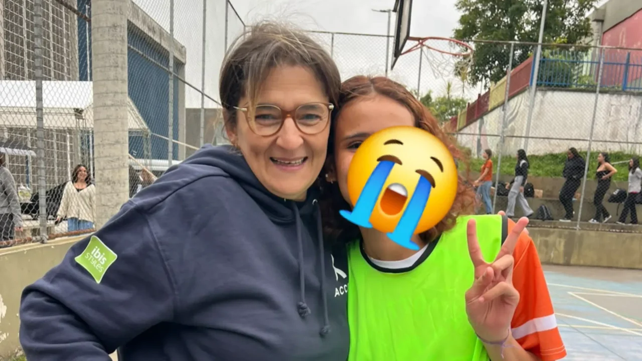 Mulher de óculos e moletom azul-marinho sorri ao lado de uma adolescente de colete verde em quadra esportiva com alambrados
