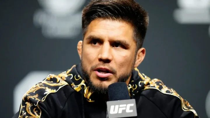 Homem de barba e jaqueta preta com detalhes dourados fala em coletiva diante de um microfone com a marca UFC, em um painel escuro ao fundo