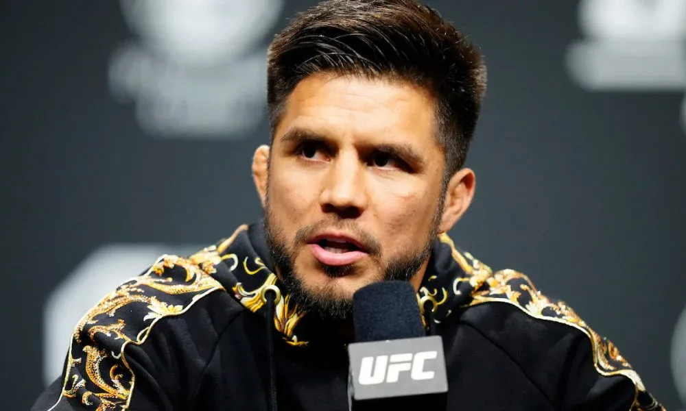 Homem de barba e jaqueta preta com detalhes dourados fala em coletiva diante de um microfone com a marca UFC, em um painel escuro ao fundo