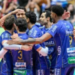 Jogadores de time de vôlei em uniforme azul se abraçam durante partida, com torcida ao fundo na quadra