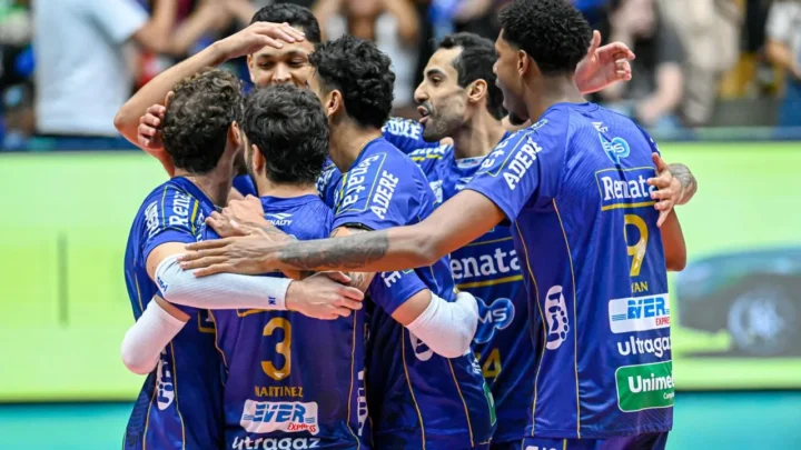 Jogadores de time de vôlei em uniforme azul se abraçam durante partida, com torcida ao fundo na quadra