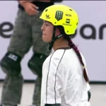 Skatista de capacete amarelo e trança observa a pista durante a semifinal do Mundial de Skate Park, com placar de competição exibido ao lado