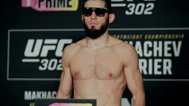 Lutador sem camisa e de óculos escuros posa de pé diante de painel do UFC 302, com parte do nome "Makhachev" visível na base