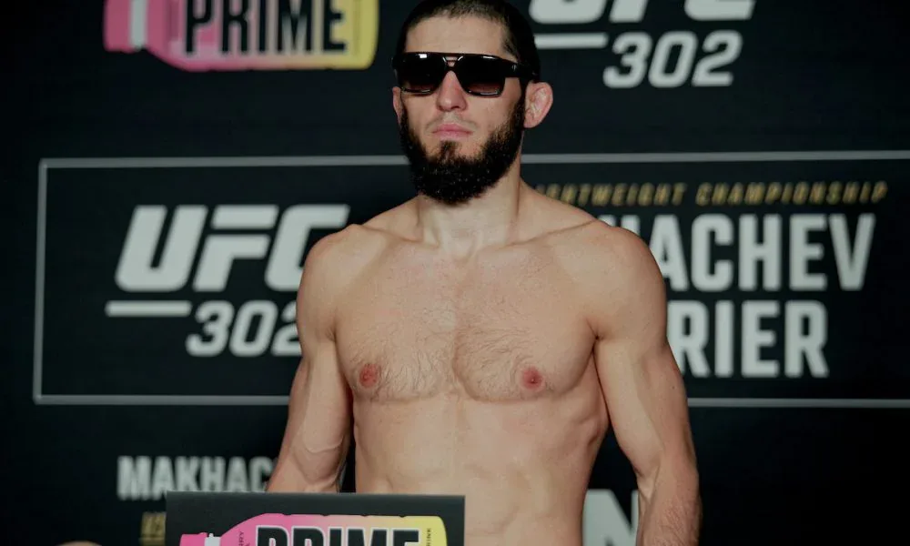 Lutador sem camisa e de óculos escuros posa de pé diante de painel do UFC 302, com parte do nome "Makhachev" visível na base