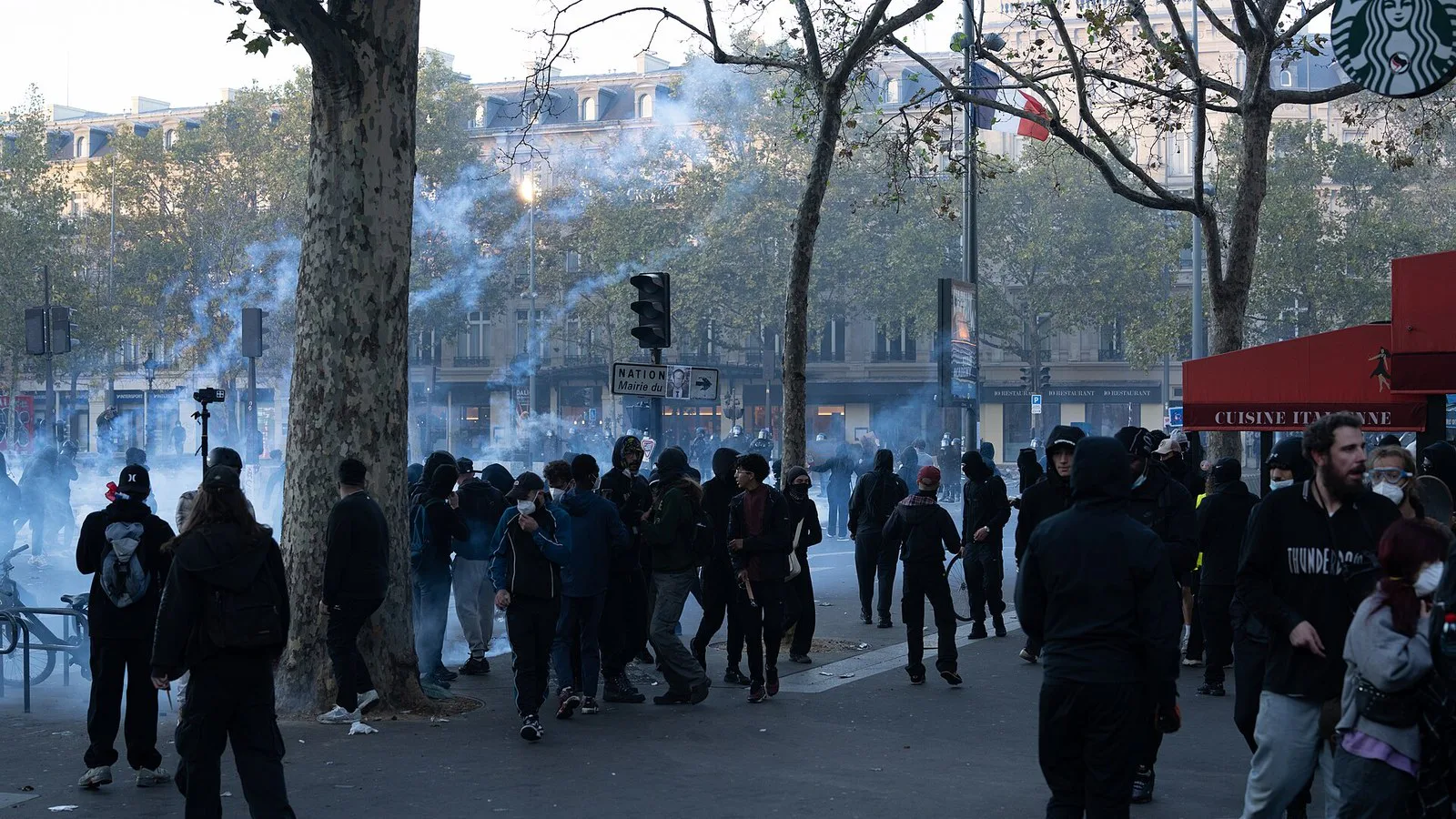 Pessoas encapuzadas correm entre árvores e nuvens de gás lacrimogêneo na Praça da República, em Paris, em 17 de outubro de 2026