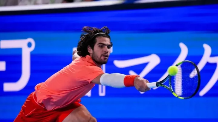 Tenista em uniforme vermelho executa golpe de forehand durante partida de tênis em quadra coberta com publicidade azul ao fundo