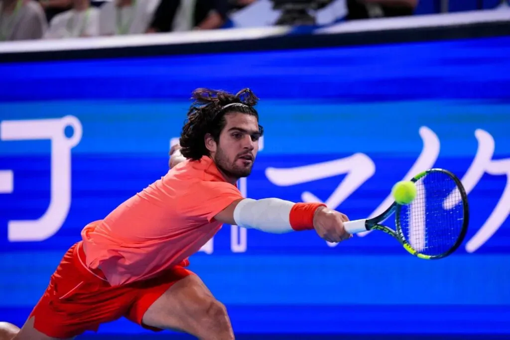 Tenista em uniforme vermelho executa golpe de forehand durante partida de tênis em quadra coberta com publicidade azul ao fundo