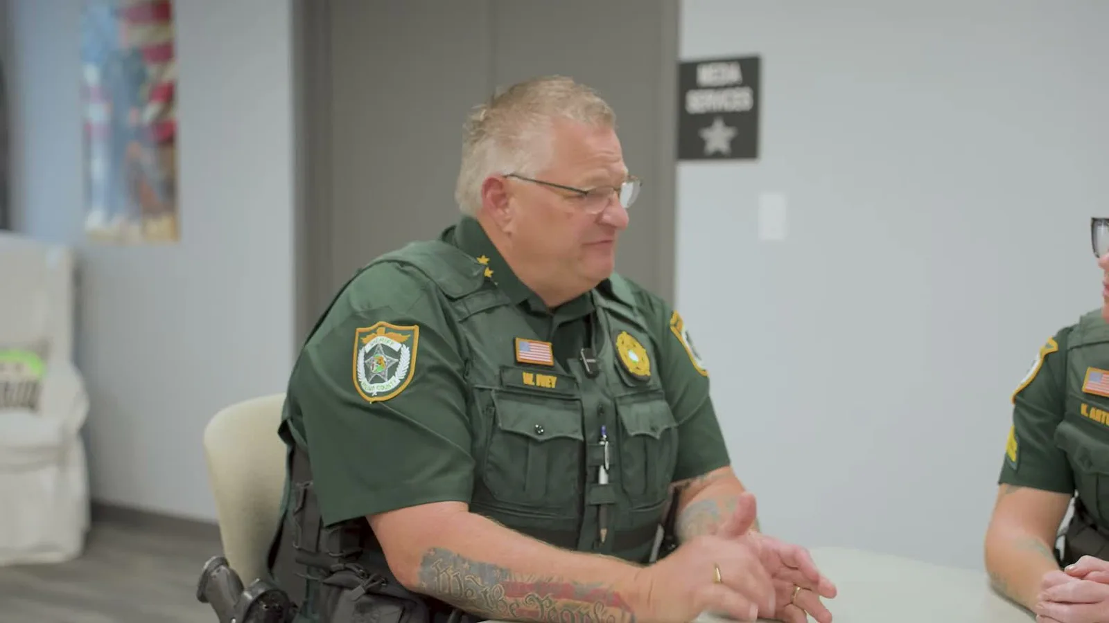 O xerife Wayne Ivey, de uniforme verde, sentado à mesa na reunião com a equipe que abre o vídeo