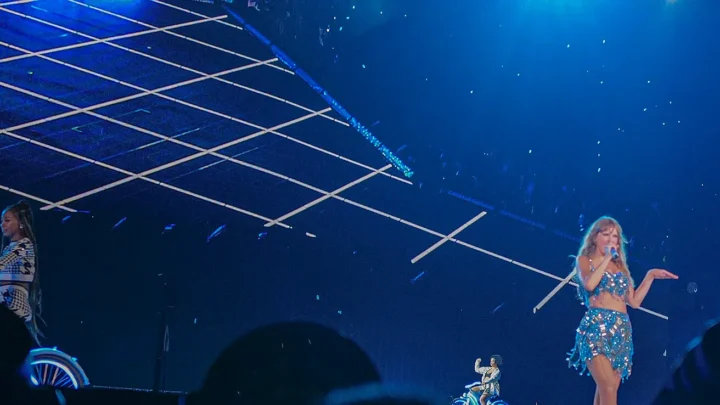 Apresentação musical em palco com iluminação azul, com artista cantando no centro usando vestido sequinado, acompanhada por músicos aos lados e estruturas geométricas de iluminação ao fundo