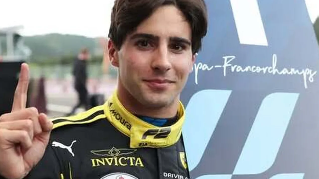 Piloto de macacão preto e amarelo com patrocínio Invicta, aponta o dedo para cima ao lado de placa da F2 com a inscrição Spa-Francorchamps