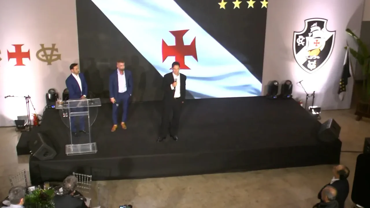 Três homens de terno em palco com painel do Vasco da Gama, um deles discursando ao microfone diante de um púlpito de acrílico