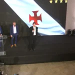 Três homens de terno em palco com painel do Vasco da Gama, um deles discursando ao microfone diante de um púlpito de acrílico