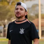 Homem de boné branco e camisa preta com o escudo do Corinthians fala em um campo de futebol, com um gramado e uma rede ao fundo