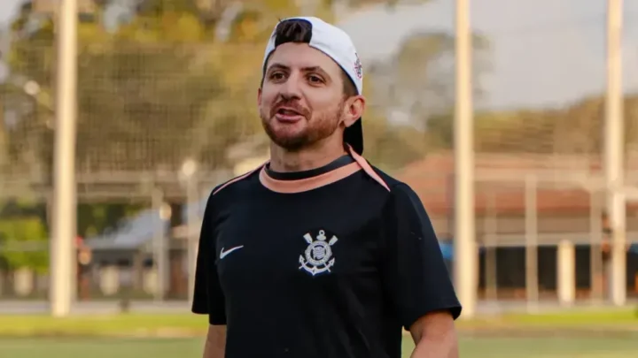 Homem de boné branco e camisa preta com o escudo do Corinthians fala em um campo de futebol, com um gramado e uma rede ao fundo