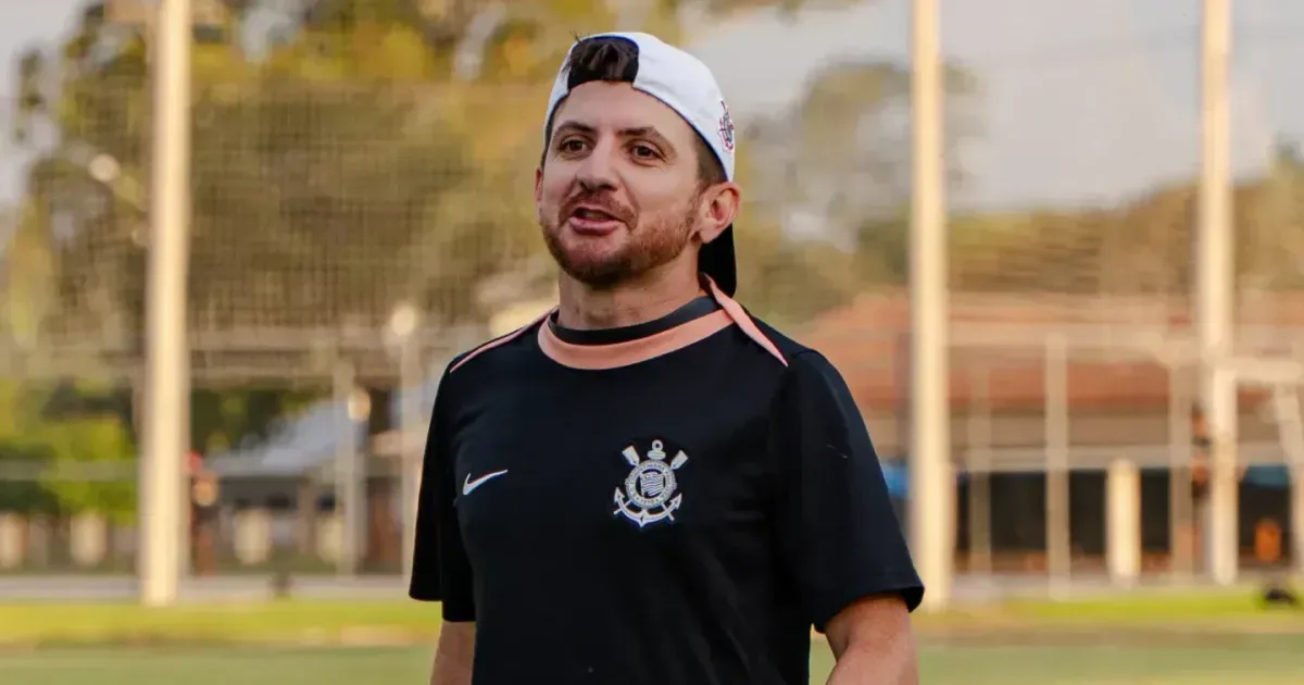 Homem de boné branco e camisa preta com o escudo do Corinthians fala em um campo de futebol, com um gramado e uma rede ao fundo