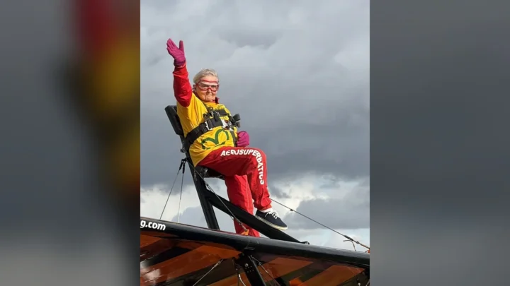 Helga Duffy, de 99 anos, acena presa à estrutura sobre a asa de um biplano, com macacão vermelho e amarelo