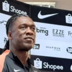 Homem de dreads, com camisa preta e laranja, posa diante de painel com logotipos de patrocinadores, incluindo Nike e Shopee