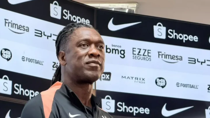 Homem de tranças e camisa preta com detalhes laranja fala à imprensa diante de painel com logos de patrocinadores, incluindo Shopee e Nike