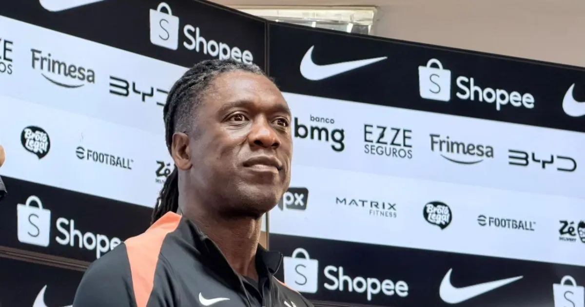 Homem de tranças e camisa preta com detalhes laranja fala à imprensa diante de painel com logos de patrocinadores, incluindo Shopee e Nike