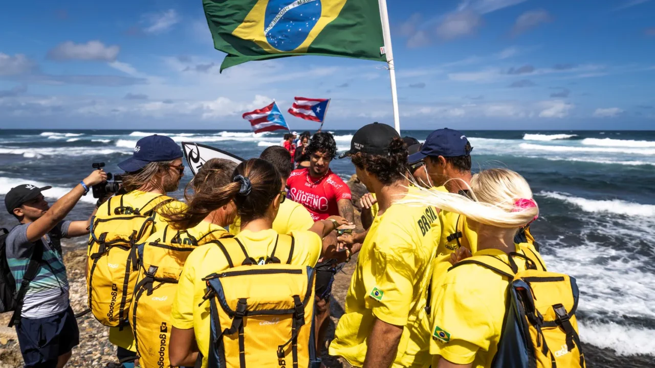 Surfistas brasileiros reunidos na praia durante competição internacional, com a bandeira do Brasil ao fundo e outras bandeiras de países participantes