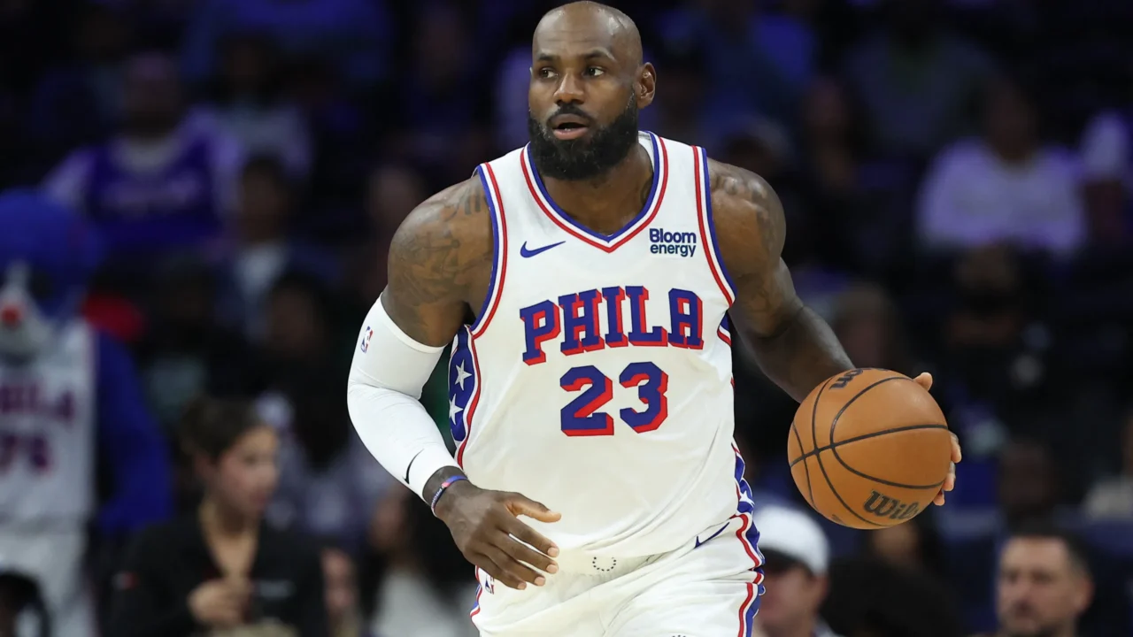Jogador de basquete de camisa branca com "PHILA" e número 23 conduz a bola em quadra durante uma partida