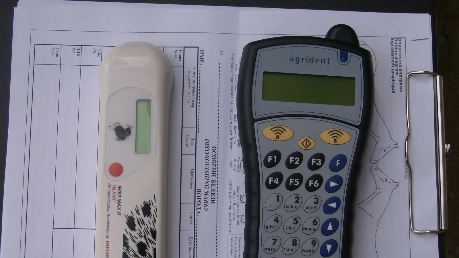 Dois leitores de microchip para animais, um branco e um cinza com teclado, sobre uma prancheta com formulário