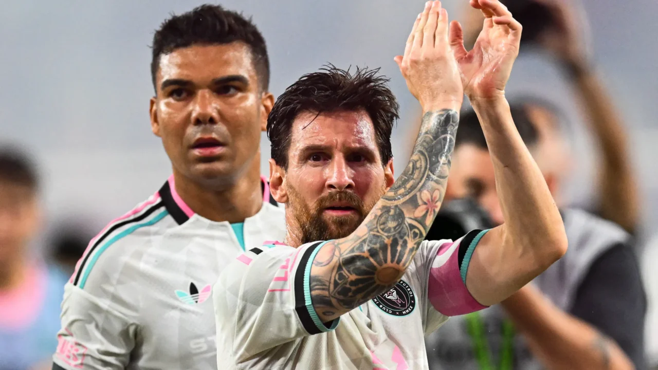 Dois jogadores de camisa branca com detalhes rosa e verde-água, um deles de braços erguidos aplaudindo a torcida, em campo