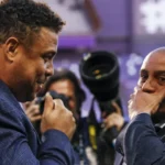 Dois homens de terno azul conversam em evento com fotógrafos ao fundo, um deles com a mão no rosto e expressão pensativa
