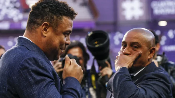 Dois homens de terno azul conversam em evento com fotógrafos ao fundo, um deles com a mão no rosto e expressão pensativa
