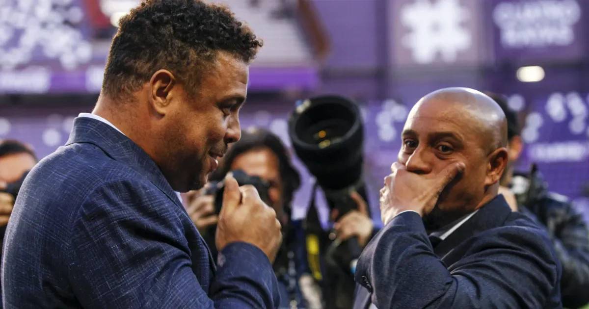 Dois homens de terno azul conversam em evento com fotógrafos ao fundo, um deles com a mão no rosto e expressão pensativa