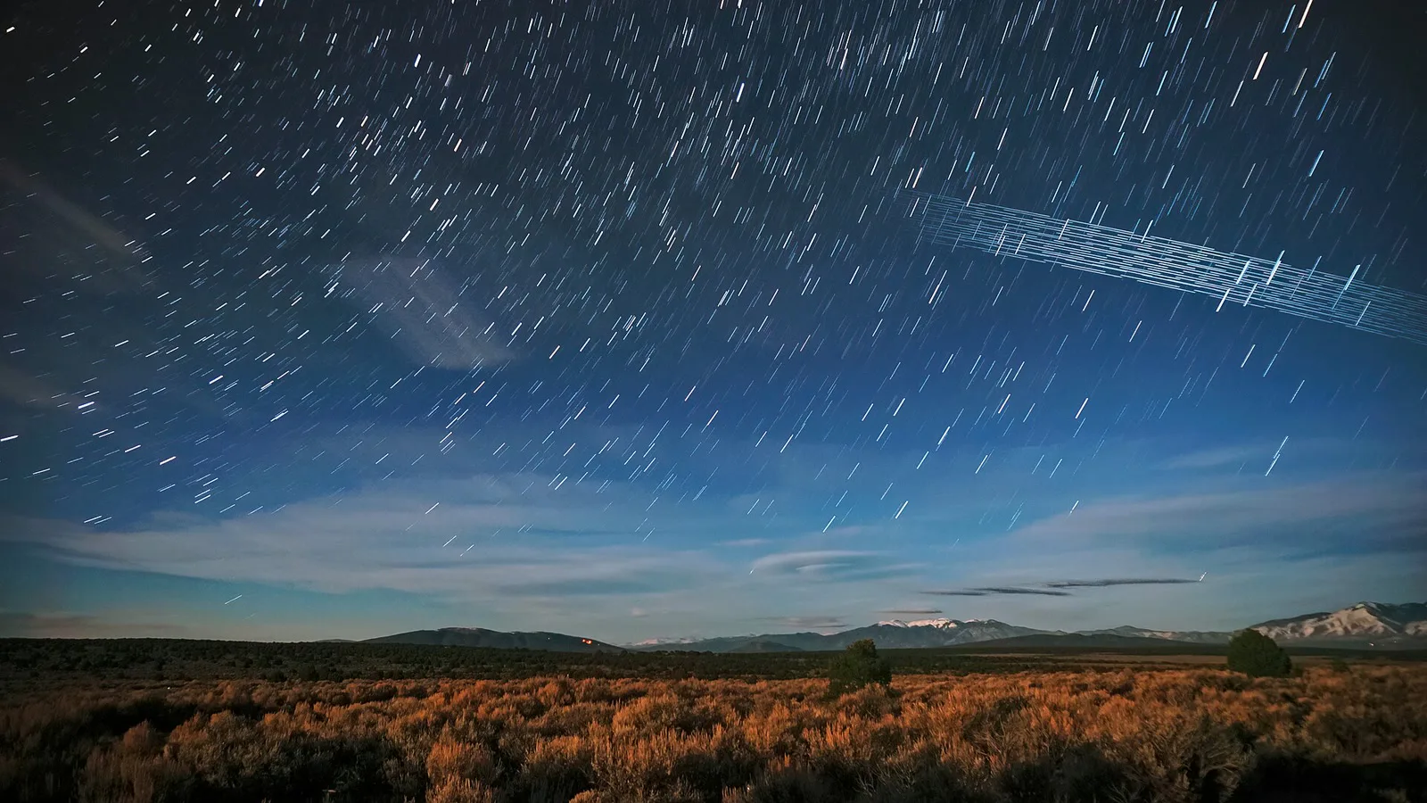 Céu noturno em longa exposição com rastros de estrelas e uma faixa de traços paralelos deixada por satélites, sobre a Floresta Nacional de Carson, no Novo México