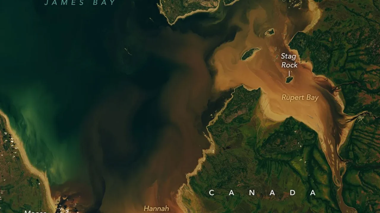 Água de um castanho escuro proveniente dos rios Nottaway, Harricanaw e Moose redemoinha nas águas azul-escuras da baía e se mistura a plumas de sedimento em suspensão de um castanho mais claro. A ilha Charlton e a bem menor Stag Rock são visíveis perto da costa.
