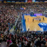 Torcedores lotam o ginásio durante partida de basquete, com placar eletrônico e jogadores em quadra