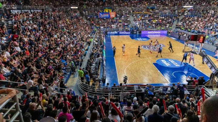 Torcedores lotam o ginásio durante partida de basquete, com placar eletrônico e jogadores em quadra