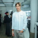 Modelo de cabelo castanho claro, com camiseta branca de manga longa e calça cinza, posa nos bastidores de um desfile, enquanto uma maquiadora secando cabelo de outra pessoa ao fundo