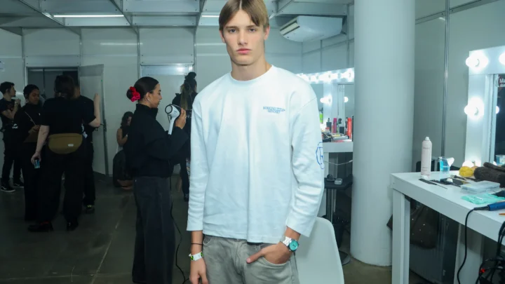 Modelo de cabelo castanho claro, com camiseta branca de manga longa e calça cinza, posa nos bastidores de um desfile, enquanto uma maquiadora secando cabelo de outra pessoa ao fundo