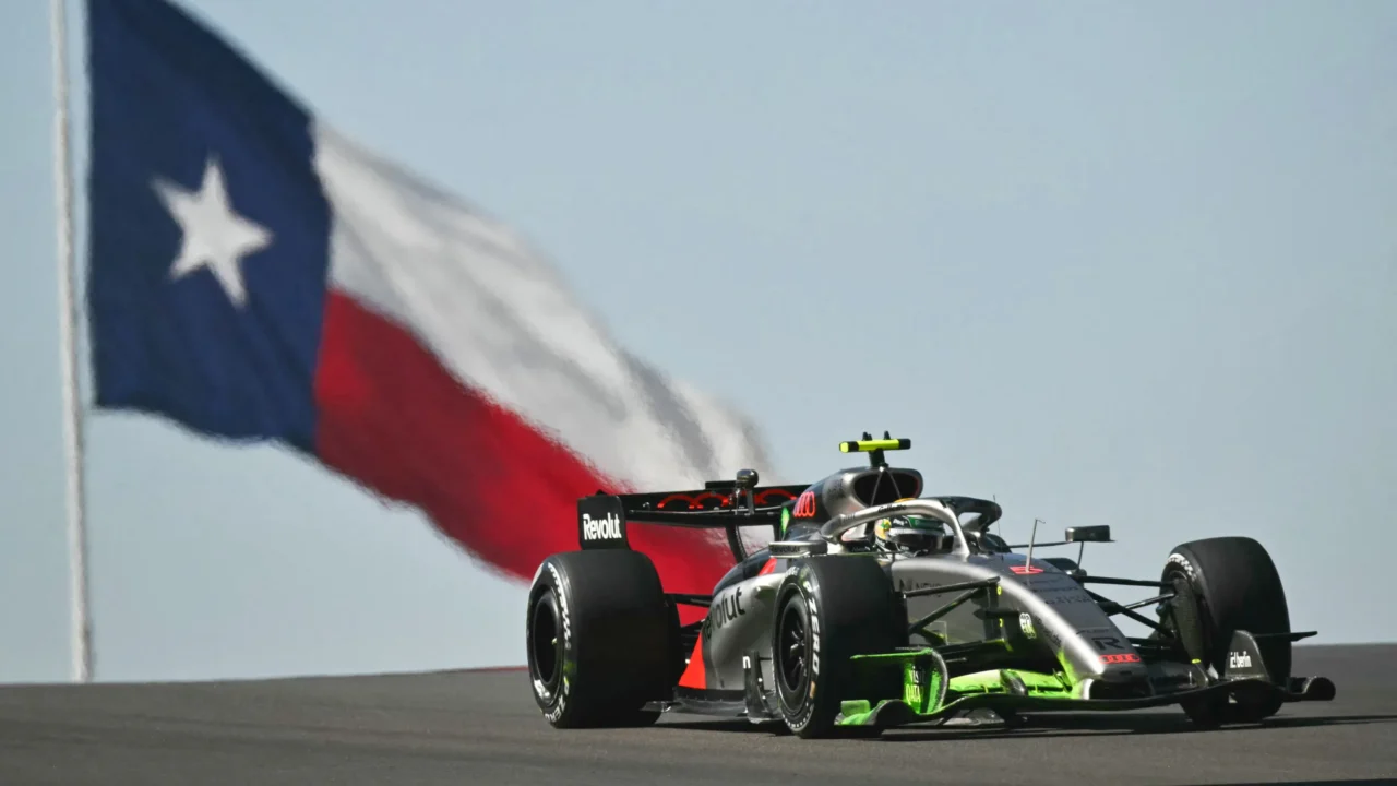 Carro prateado com detalhes verdes e vermelhos da Audi passa em alta velocidade diante de uma bandeira do Texas tremulando ao vento