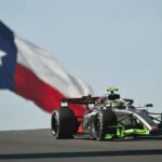 Carro prateado com detalhes verdes e vermelhos da Audi passa em alta velocidade diante de uma bandeira do Texas tremulando ao vento