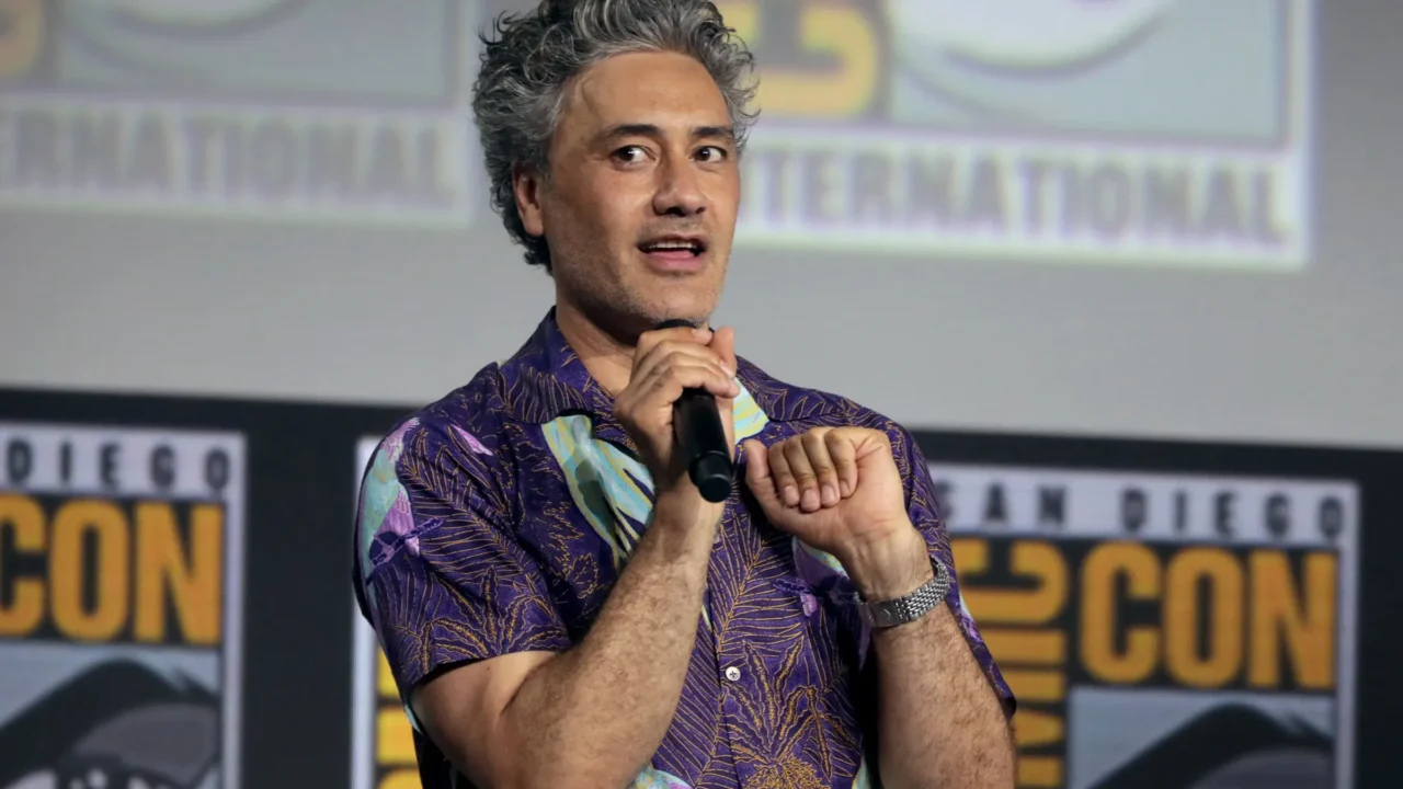 Homem de cabelo grisalho, camisa roxa de estampa tropical, segura um microfone e fala em palco com telões do Comic-Con International de San Diego ao fundo