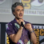 Homem de cabelo grisalho, camisa roxa de estampa tropical, segura um microfone e fala em palco com telões do Comic-Con International de San Diego ao fundo