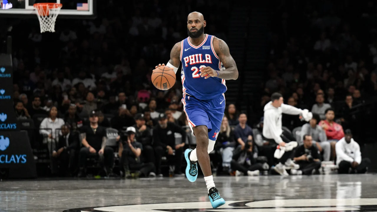 Jogador de camisa azul do Philadelphia 76ers, de número 23, conduz a bola em quadra de arena com torcida ao fundo