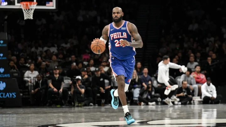 Jogador de camisa azul do Philadelphia 76ers, de número 23, conduz a bola em quadra de arena com torcida ao fundo